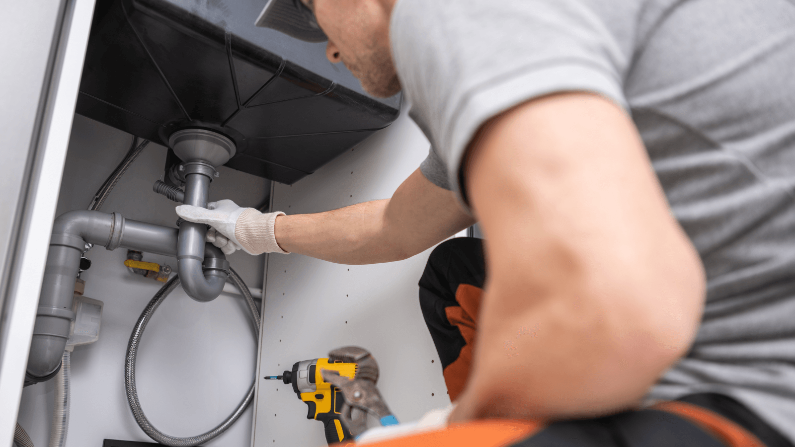 A plumber inspecting and repairing a sink drainage system under a kitchen sink, wearing gloves and using tools for maintenance.